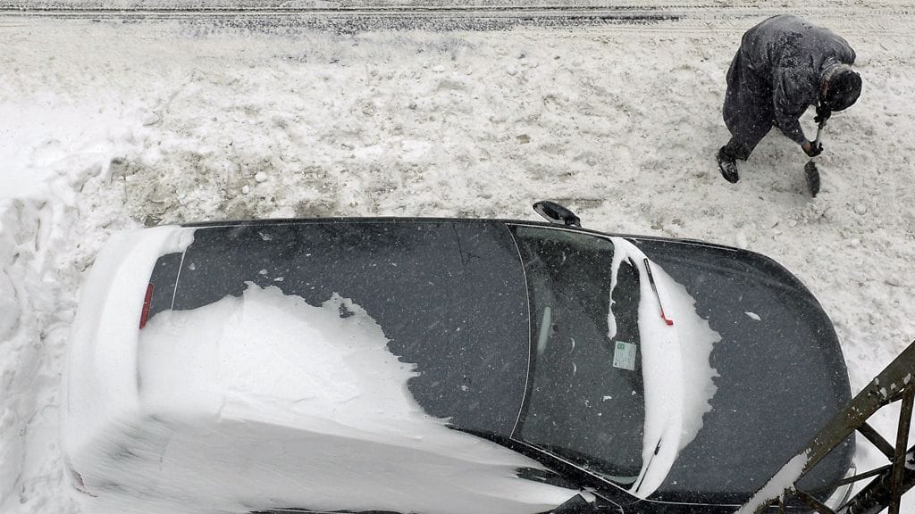 Mies kaivaa lunta jumittuneen auton ympäriltä. Kuvituskuva.