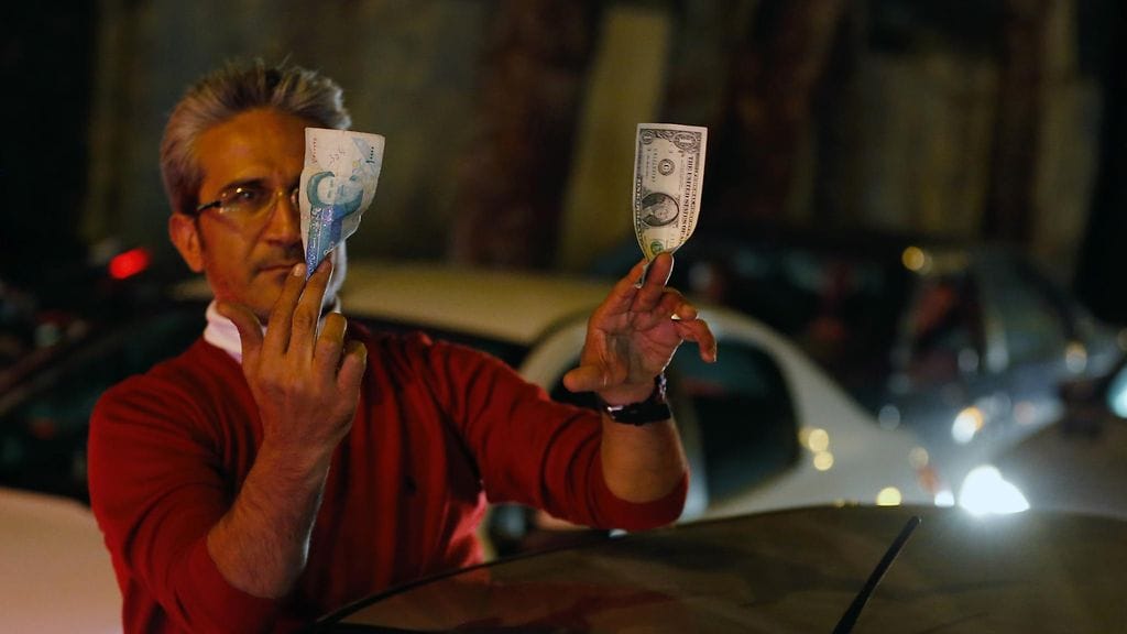 Miehellä on oikeassa kädessään tuhannen rialin seteli ja vasemmassa kädessään dollarin seteli.