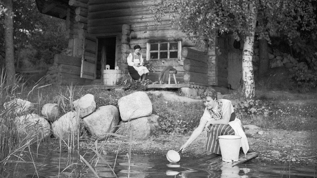 Kansallispukuiset naiset lämmittävät hirsisaunaa ja tekevät vastoja vuonna 1953.