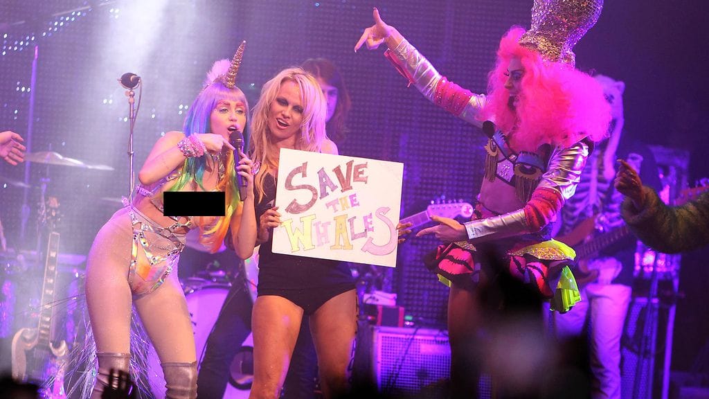 Miley Cyrus (vas.) ja Pamela Anderson. Kuvasta peitetty Cyrusin asuun kuuluvat tekorinnat.