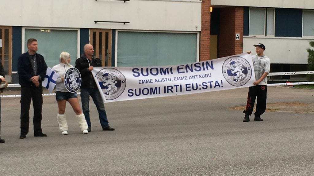 Mielenosoittajat järjestäytyivät kadulle. Taustalla vastaanottokeskus, jonka sälekaihtimet pysyivät suljettuna ja turvapaikanhakijoita ei näkynyt.