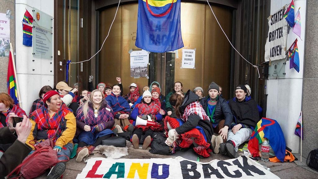 Mielenosoittajat istuvat ovien edessä isona ryhmänä saamelaisliput ja muut lakanatliehuen.