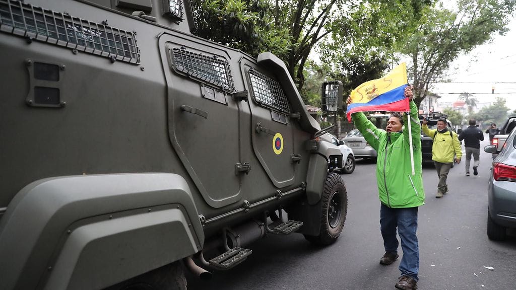 Mies näyttää kansallislippua panssaroidun ajoneuvon edessä, jonka väitetään kuljettavan Ecuadorin entistä varapresidenttiä Jorge Glasia lentokentälle Ecuadorin Quitossa