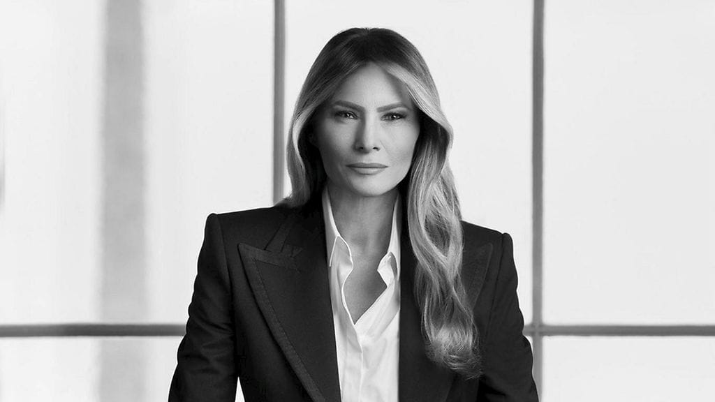 Melania Trumpilla on yllään tumma puvuntakki ja housut sekä vaalea kauluspaita, ja hän poseeraa kuvassa seisten, työpöytään nojaten.