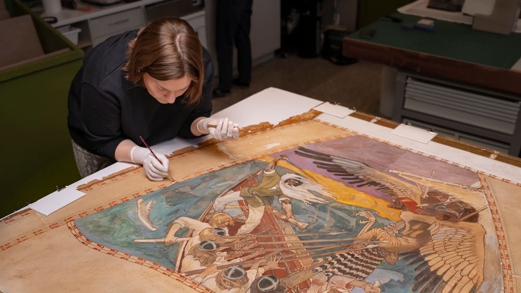 Paperikonservaattori Ilona Osara sävyttää Sammon puolustus -freskoluonnoksen japaninpaperilla paikattuja alueita vesiliukoisilla väreillä.