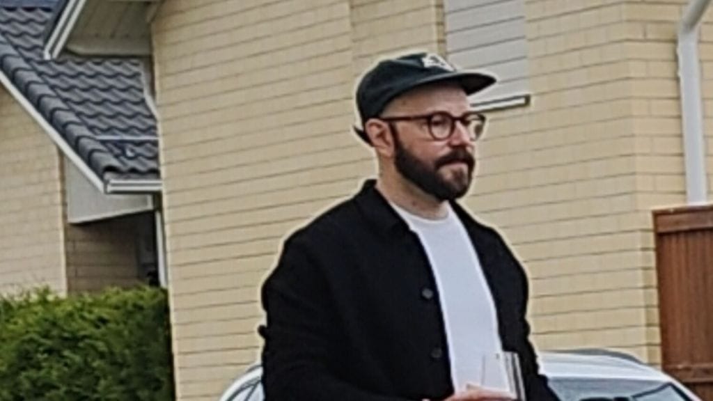 Miehellä lyhyet hiukset, tumma kokoparta, pyöreät ja tummasankaiset silmälasit sekä tatuointeja säärissä ja käsissä.