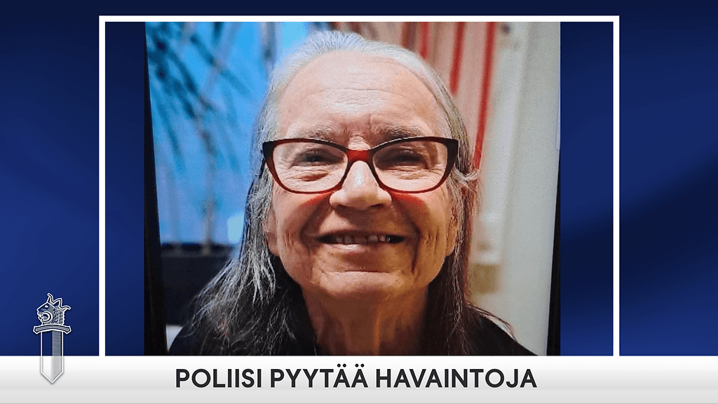 Naisella on tummanharmaat olkapäille ulottuvat hiukset, punaiset silmälasit, ja päällä tummat vaatteet.